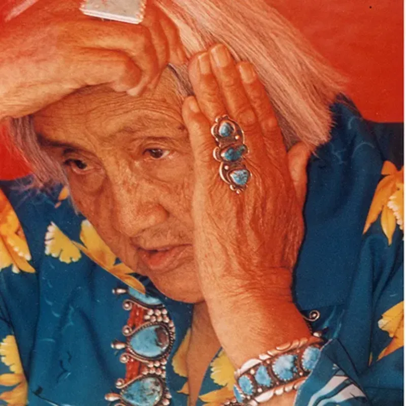 Color photograph of an older woman with short, white hair. Her medium-dark skin is lined, and she reaches her hands up to touch her hair. She wears large turqouise jewelry and a blue shirt with large yellow blossoms.