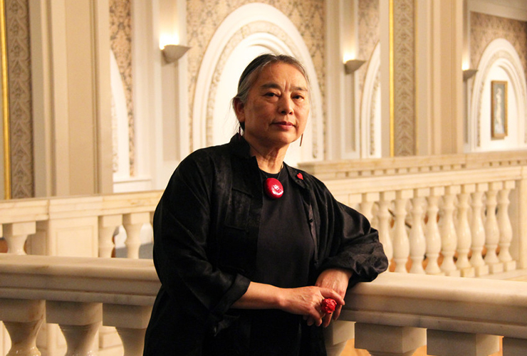 A light-medium-skinned older woman leans her elbow against an interior marble balustrade. Her grey and black hair is pulled back, and she wears a black long-sleeve top and drop earrings, a red necklace, a red ring, and a red heart-shaped lapel pin.