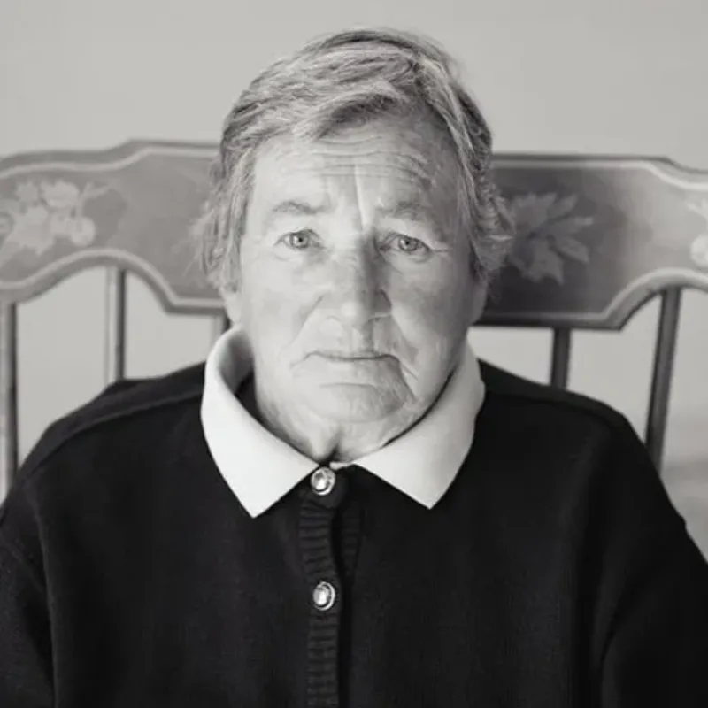 A black-and-white photograph of a light-skinned, older woman gazing head-on at the viewer. She has short, light colored hair; wears a dark, buttoned-up cardigan with the wings of a white collar peeking out, and the high-backed chair she sits in is decorated with leaf patterns.