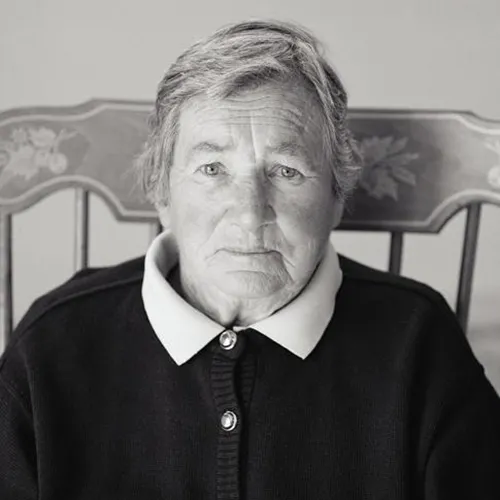 A black-and-white photograph of a light-skinned, older woman gazing head-on at the viewer. She has short, light colored hair; wears a dark, buttoned-up cardigan with the wings of a white collar peeking out, and the high-backed chair she sits in is decorated with leaf patterns.