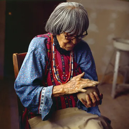 A medium-skinned, older woman working a ball of clay that she holds in her hands. She has grey hair cut just below the ear, medium skin tone and wears a blue dress and beaded necklaces of white, blue, red and silver. She is engrossed in the clay, her movements practiced.