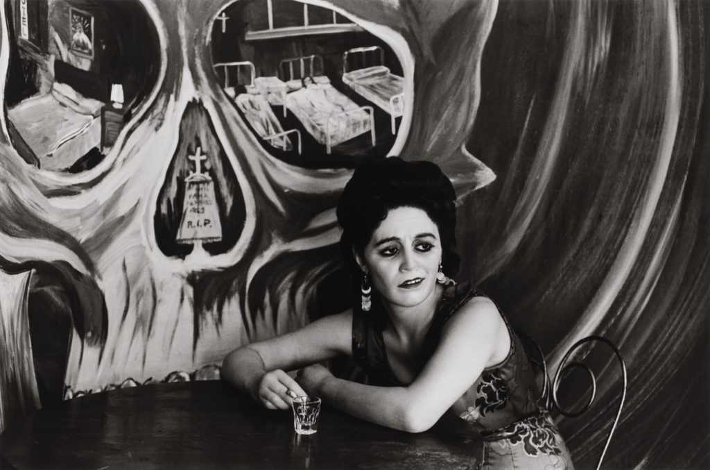 A light-skinned woman with heavy eye makeup leans over a round table, holding a cigarette next to an empty clear shot glass. A skull is painted in sweeping brushstrokes on the wall behind her, with a tombstone in the nasal cavity and hospital beds in the eye cavities.