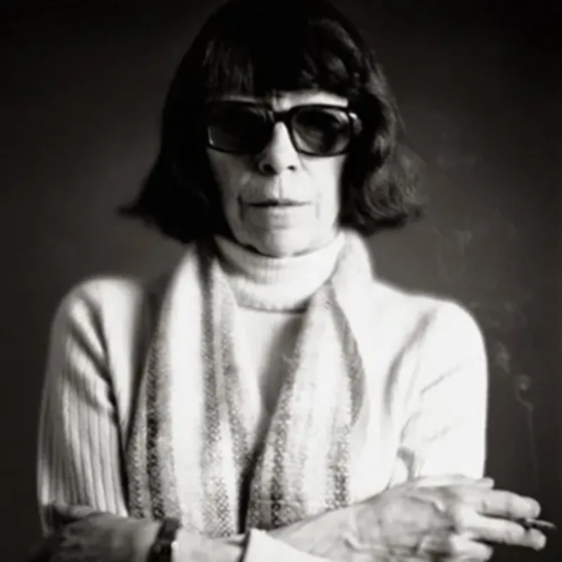 A black-and-white studio portrait of a light-skinned older woman with dark bobbed hair. She wears sunglasses, a light-colored turtleneck sweater, a striped scarf, and a watch. She stands with arms folded and holding a lit cigarette between her left index and middle fingers.