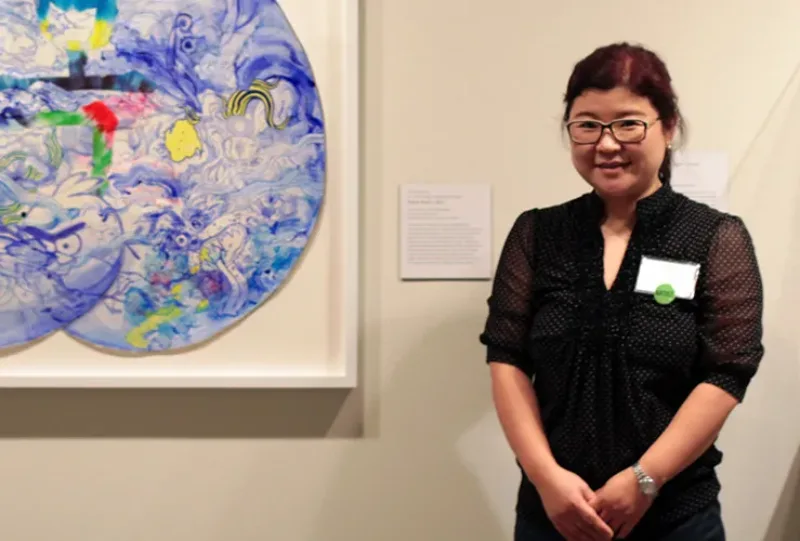 A medium-light-skinned smiling adult woman with dark hair in a ponytail, clasps her hands in front of her. She wears a black dress with a name tag, glasses, and watch. On the white wall behind her is a colorful abstract painting and a label with unreadable text.