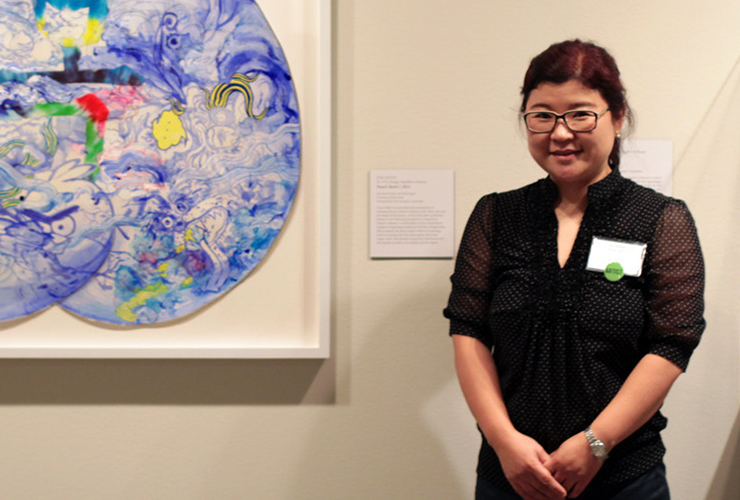 A medium-light-skinned smiling adult woman with dark hair in a ponytail, clasps her hands in front of her. She wears a black dress with a name tag, glasses, and watch. On the white wall behind her is a colorful abstract painting and a label with unreadable text.