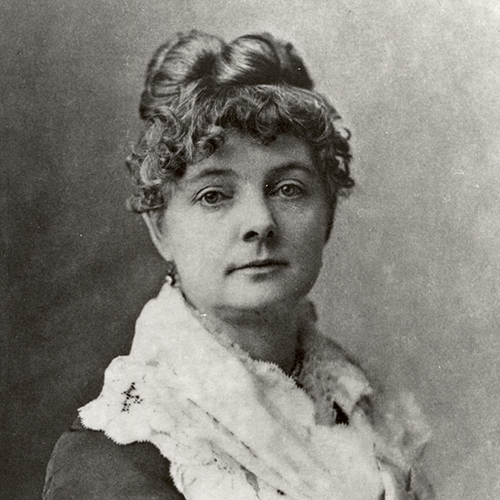 Black and white portrait of a woman with light skin. Seated and staring straight at the camera, she wears her hair up, with curls framing her face. Around her shoulders she wears a white shawl, and has a delicate earring in her right ear. Her eyes are clear and she smiles slightly.