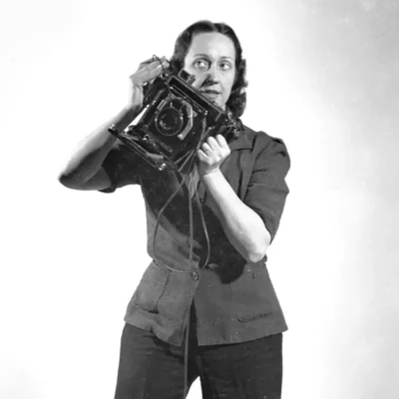 A black-and-white photograph of a light-skinned adult woman standing against a white backdrop and holding a large, antique camera in her hands at an angle. She gazes to the side and has dark, wavy hair and wears a collared, buttoned jacket and slacks.