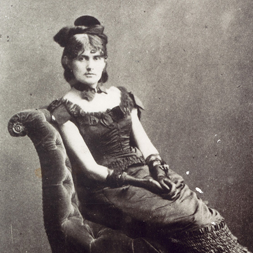 A black-and-white photograph of a light-skinned adult woman sitting upright in a luxurious velvet chair. Her dark hair is meticulously curled, and she wears a fascinator, a collar necklace with a bow, long gloves, and a long, sleeveless dress embellished with many frills.