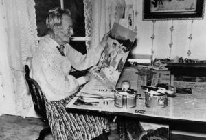 A black-and-white photograph of an older woman sitting at a table in front of floral wallpaper and a window with lace curtains. She holds up a canvas depicting houses and snow in her left hand. In her right hand, she holds a small paintbrush to the canvas.