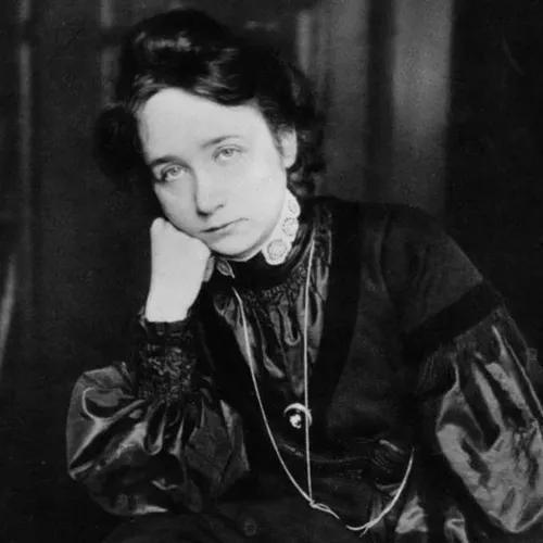 A black-and-white photograph of a light-skinned adult woman with dark hair pulled back from her face. She is seated with her arm on her leg and face resting on her fist. She wears a dark collared dress with puffed sleeves and a long necklace, and makes eye contact with the viewer.