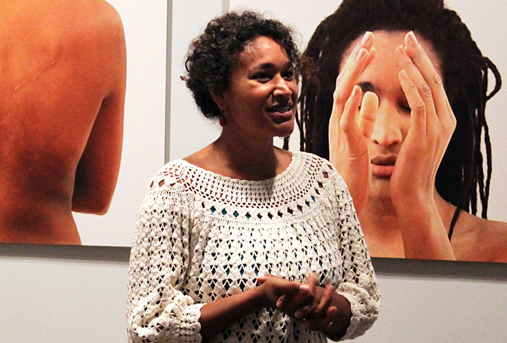 A medium-dark-skinned adult woman with short dark curly hair, wearing a white crochet top, stands with hands clasped in front of her. On the wall behind her are two large color photographs of medium-skinned adult women.