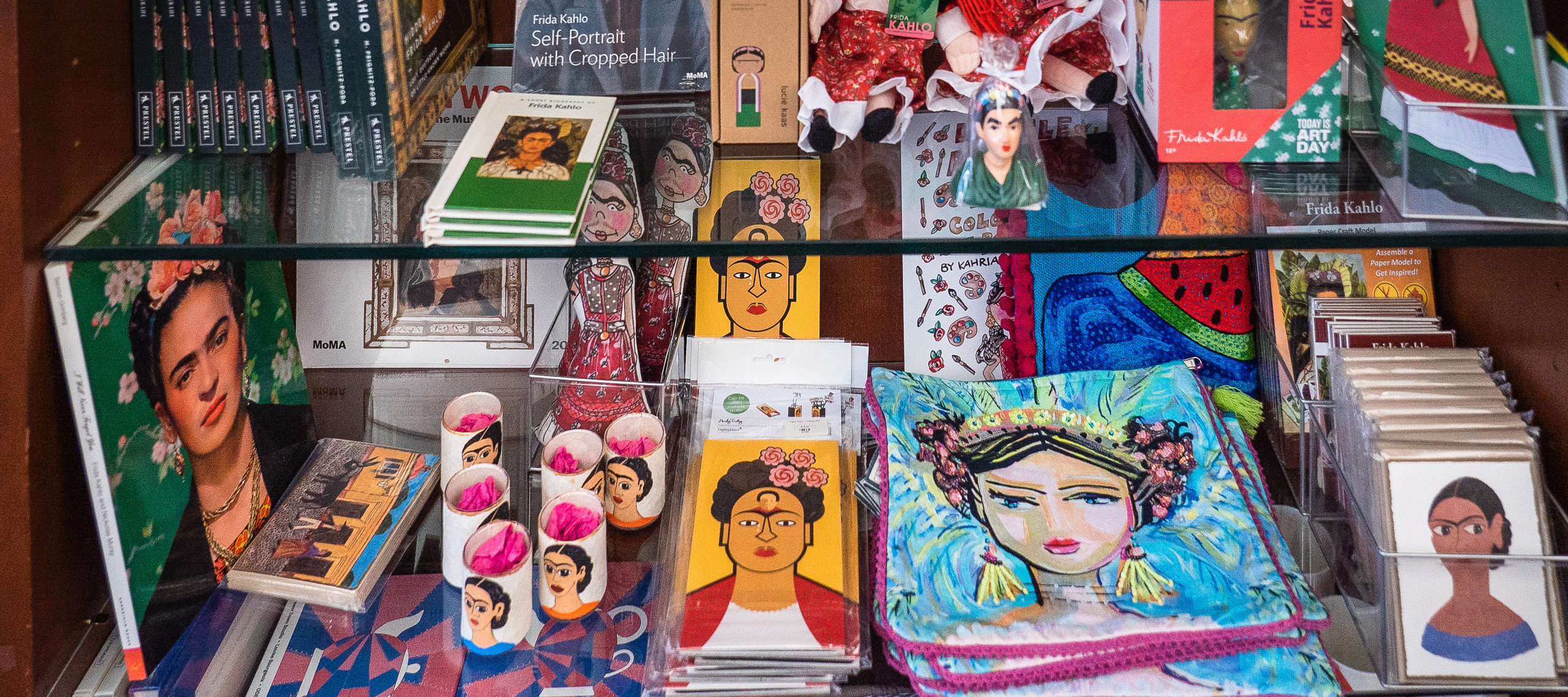 Three clear glass shelves, packed with colorful products on display. The top two shelves are full of Frida Kahlo-themed items—books, dolls, cards, and more. The bottom shelf is full of Louise Bourgeois-themed items—teapots, a sleep mask, books, mugs, and more.