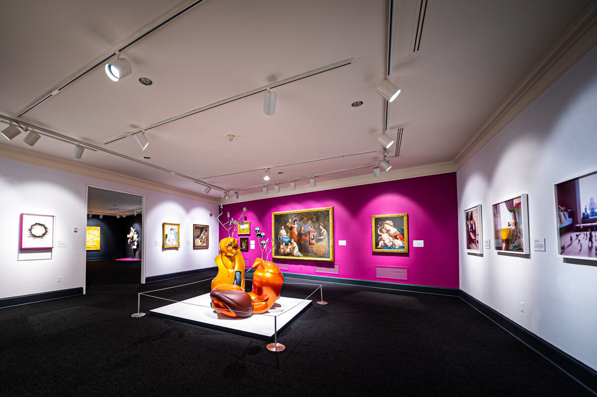 Gallery view with contemporary sculpture made of orange motor scooters that resemble two antlered animals fighting in foreground. Four paintings hang on a magenta wall in background, including a baroque painting of the Virgin and Child, and a portrait of a light-skinned man, women and children gathered together in eighteenth-century attire. To the left on a white wall hang two other paintings.