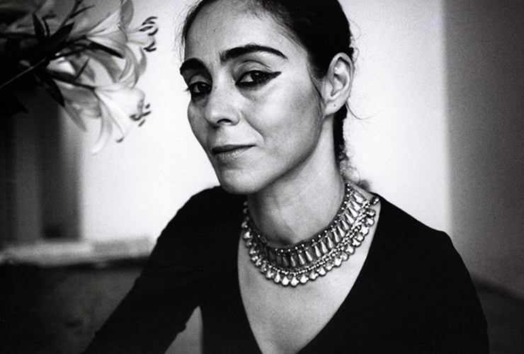 Black and white photo of a woman sitting next to a vase of lillies. Her eyes are lined in thick black pencil, which matches her dark brows. Her tiered metal necklace and dark v-neck shirt accenuate the skin of her chest. Her dark hair is pulled back and she smiles slightly.