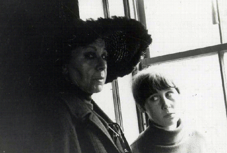 Black and white photo of two women by a window. One wears a black hat with a large rim and gazes at the camera. She looms over the younger woman who sits facing forward while she side-eyes the woman in the hat. The energy is tense, as if the younger woman is in trouble.
