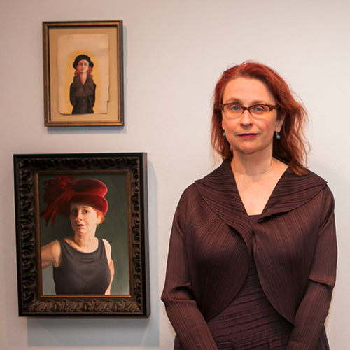 A light-skinned adult woman smiles while standing against a white wall beside two paintings that resemble her in theatrical costumes. She has long red hair, and wears glasses, earrings, and a brown jacket over a matching brown dress with subtle, organic stripes on them.