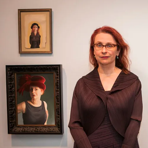 A light-skinned adult woman smiles while standing against a white wall beside two paintings that resemble her in theatrical costumes. She has long red hair, and wears glasses, earrings, and a brown jacket over a matching brown dress with subtle, organic stripes on them.