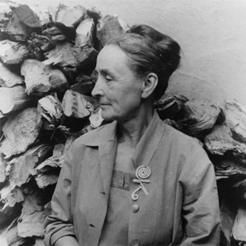 A black-and-white photograph of a light-skinned older woman. Standing in front of stacked wood, she is posed from the waist up with her head turned to the left in profile. She has grey hair pulled up into a large bun. She wears a blazer with a large abstract brooch.