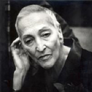 Black and white photo of a woman shot from the shoulders up. She leans to the left, bringing her hand to her ear. Her light hair is cropped very short to her head and she looks past the camera with a thoughful expression, looking as if she were about to begin speaking.