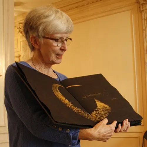 An older light-skinned woman with short white hair wears glasses and a long-sleeve blue top. She stands inside, holding open a large black book with an abstract gold design printed across two pages and unreadable gold writing.