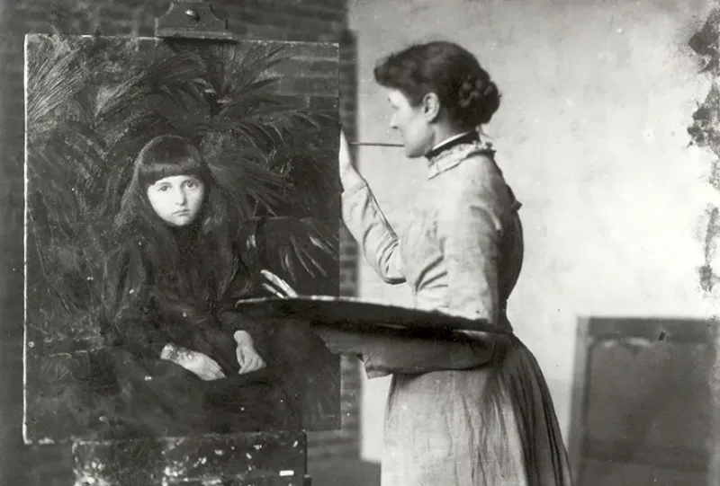 Black-and-white interior photograph of a light-skinned adult woman in profile with dark hair in a bun and a light-color dress. She stands in front of an easel and holds a palatte and brushes in her left hand. She rests her right hand on a painting of a light-skinned young girl.