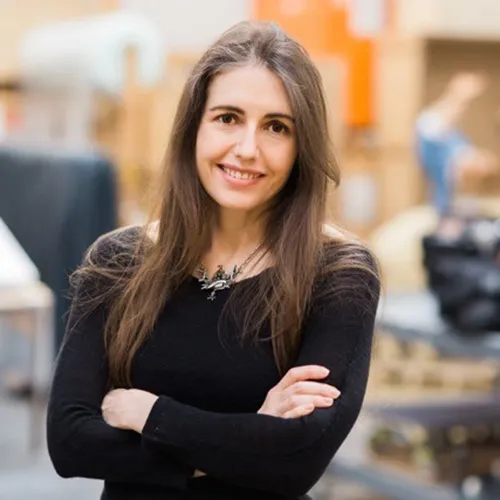 A color photograph of Patricia Piccinini. The light-skinned woman stands with her arms folded in front of her, smiling, wearing a dark sweater. Her brunette hair falls softly to her chest. She wears an ornate silver-colored necklace with jewel-colored ornaments.