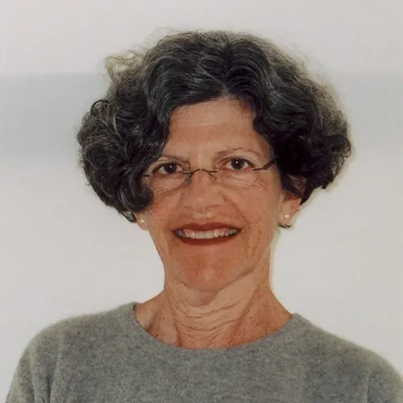 An older light-skinned woman with short, dark, curly hair looks directly at the viewer. Posed from the chest up against a light grey background, she wears a darker grey sweater, eye glasses, pearl earrings, and lipstick.