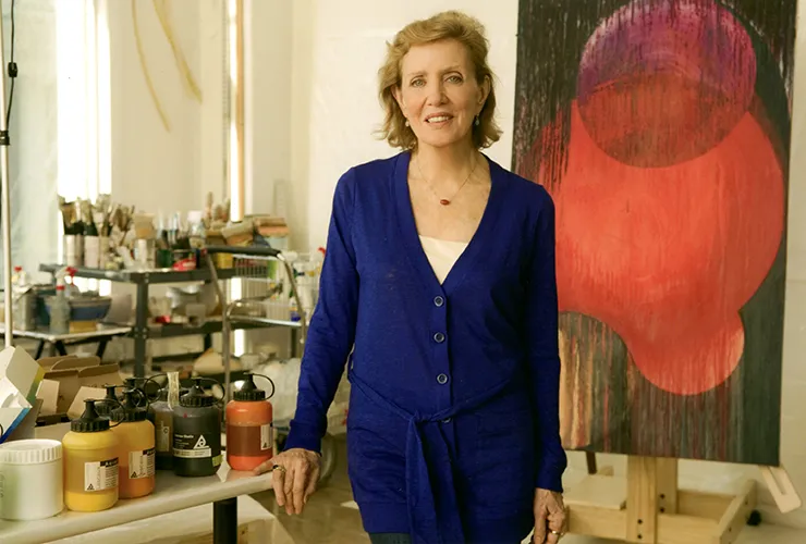 A light-skinned older woman stands in the middle of an art studio. She has short, light colored hair and wears a long blue cardigan. Next to her is a table with paint bottles on it. Behind her are carts with paint brushes and art supplies, and a large abstract red and black painting on an easel.