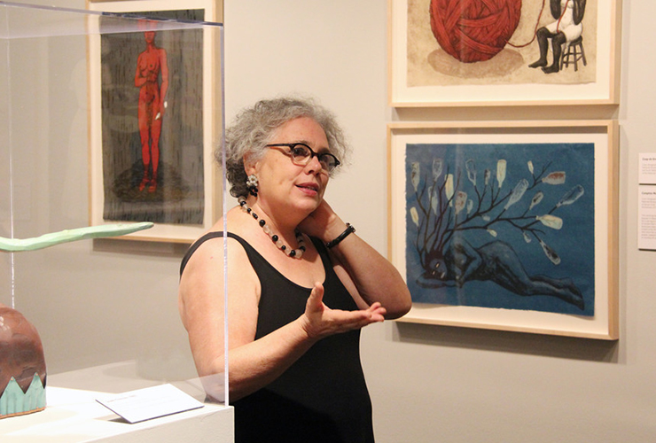 A light-skinned, adult woman speaking with one hand extended and the other touching her neck in a white gallery filled with her colorful artworks depicting African-American women. She has curly gray hair, glasses, and wears a simple, black, sleeveless top and a necklace.