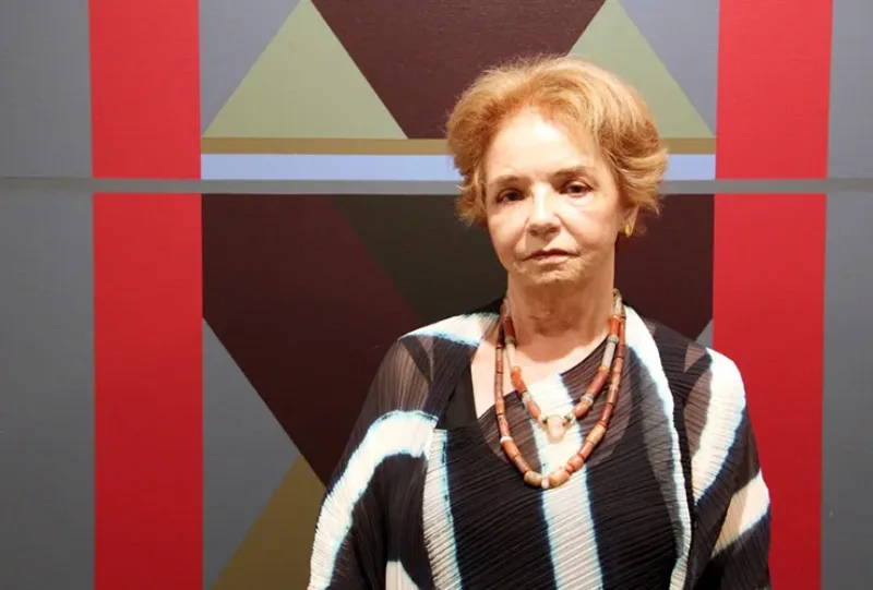 A light-skinned older woman with short, light brown hair stands in front of an abstract painting. She wears a black and white striped shirt with two beaded necklaces. The painting behind her depicts grey and red rectangles framing grey, brown, and green triangles.