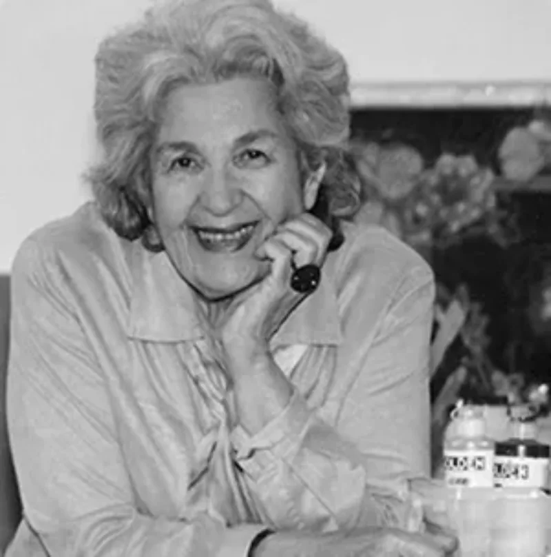 Black and white photo of a woman shot from the waist up. She rests her chin in her right hand, her right elbow propped on a table next to bottles of paint. She smiles with her whole face and her grey hair is brushed back. She wears a light blouse and large cocktail ring.