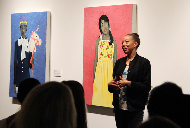 Amy Sherald, a medium skin-toned adult woman, stands and speaks to a crowd in a gallery beside her striking portraits depicting individuals with gray skintones against vibrant, solid-colored backgrounds. She smiles, her dark hair pulled tightly into a bun, and wears mostly black.