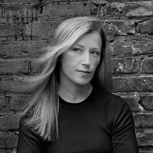 A black-and-white photograph of a light-skinned adult woman with medium-length straight blonde hair. She wears a dark colored long sleeved shirt. Shown from her mid-section up, she stands in front of a brick wall and glances to the side.