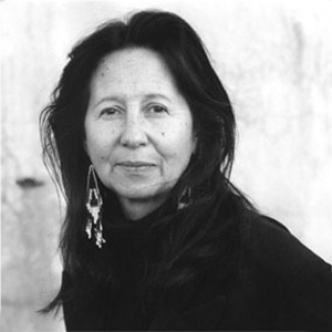 A black-and-white photograph of a light-skinned adult woman with long dark hair, shown in full face from the chest up, standing in front of a light-colored wall. She wears large earrings and a black turtleneck top.