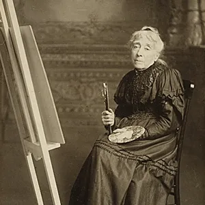 Black-and-white studio portrait of a light-skinned older woman white hair pulled back. She sits in a chair next to an easel and holds a palatte and brushes. She wears a high-neck, long-sleeve, dark-color long dress.