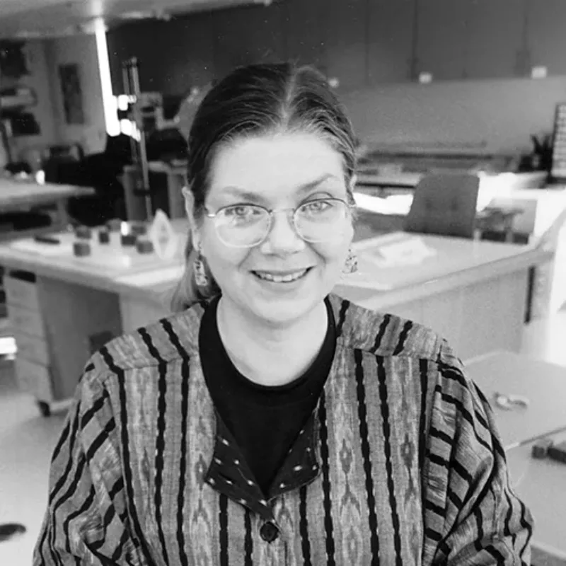 A black-and-white photograph of a smiling Pamela Spitzmueller, a light-skinned woman with eyeglasses, in a conservation lab. Her hair is parted in the middle and pulled back, revealing dangling earrings. She wears an ikat-woven jacket.