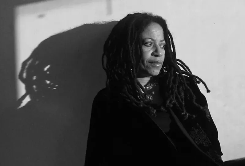 " A black-and-white photograph of Renee Stout. With medium-brown skin and thick dreadlocks, and clad in a dark jacket, she looks away from the camera as she leans against a wall. Her figure casts a looming shadow against the wall."