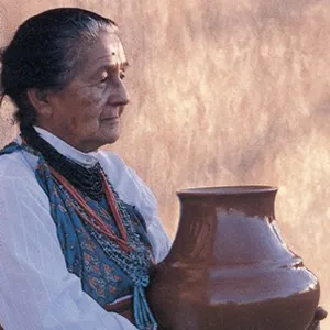 Color photo of a woman holding a large glazed pot. Seated against a sunlit wall, her dark hair is pulled back and she wears a white blouse with a color handkerchief and layers of necklaces with blue, red and black beads. She gazes into the distance, a calm expression on her face.