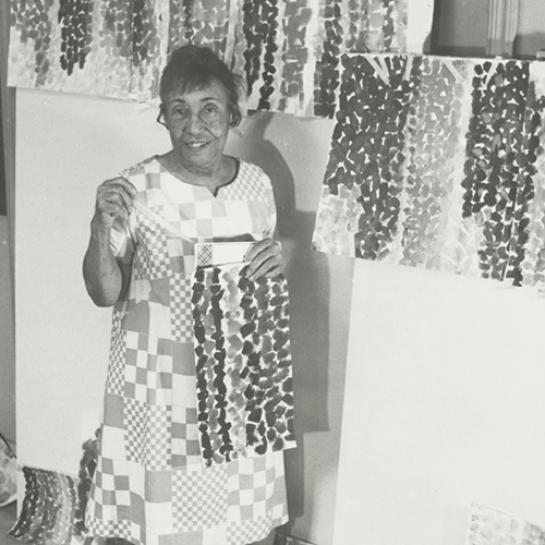 A black-and-white photograph of an older adult woman wearing a dress with square patterns resembling a quilt. She smiles, surrounded by her paintings, holding one herself. The paintings feature different colored, thick, similarly shaped brushstrokes that create vertical stripes.