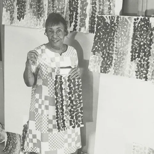 A black-and-white photograph of an older adult woman wearing a dress with square patterns resembling a quilt. She smiles, surrounded by her paintings, holding one herself. The paintings feature different colored, thick, similarly shaped brushstrokes that create vertical stripes.