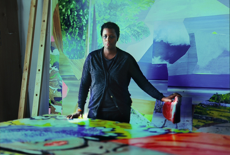 A woman with a dark skin tone and short black hair stands behind a large table covered in a painting. Her dark clothing stands out against the rich blues, greens, reds and yellows of the room she is in. Her left hand rests on a stool covered in red paint.