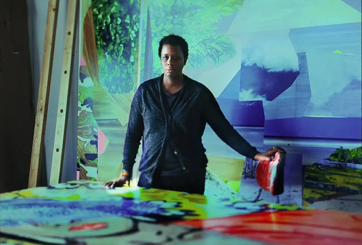 A woman with a dark skin tone and short black hair stands behind a large table covered in a painting. Her dark clothing stands out against the rich blues, greens, reds and yellows of the room she is in. Her left hand rests on a stool covered in red paint.