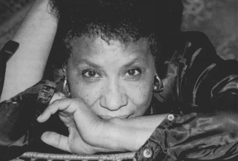 Close-up black and white photo of a woman with a medium-dark skin tone looking directly into the camera. Resting her head on a table, she crosses her left arm in front of her mouth, reaching her right hand up to her hair. She wears a dark blouse, open at the cuffs, gold hoop earrings.