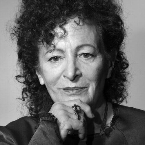 Black-and-white photograph of the artist Nan Goldin, who has light skin tone, dark curly hair that frames her face. She is resting her right hand on her chin as she looks head on.