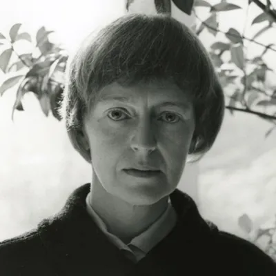 A black-and-white photograph of a light-skinned adult woman looking straight-on at the viewer. She sports a bowl cut with bangs cut significantly above her brow, and a collared shirt under a dark colored jacket. Behind her are delicate branches with leaves.