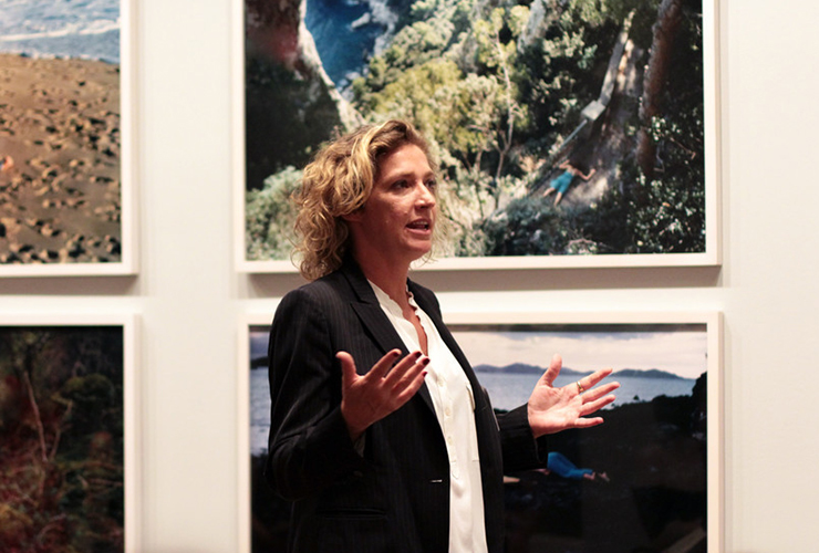 A light-skinned adult woman with medium-length curly blond hair wears a black pinstripe blazer over a white blouse. Her mouth is open in speech and arms bent in front of her. Four large color landscape photographs are on the white wall behind her.