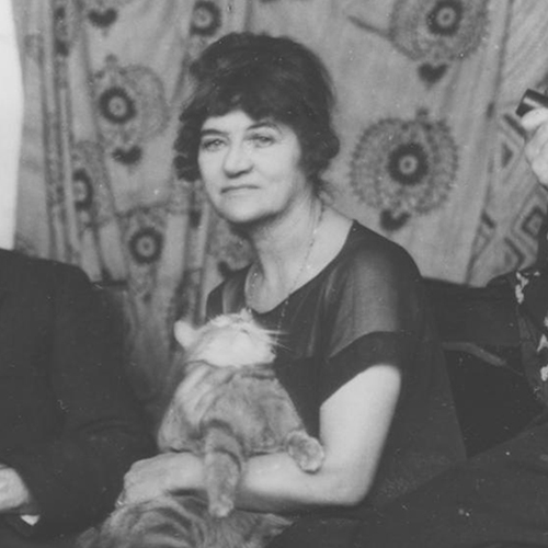 Black and white photo of a light-skinned woman who has her dark hair piled on top of her head. She slouches slightly, holding a struggilng tabby cat whose movement blurs in the image. She wears a black dress and sits in front of hung textiles, decorated in geometric patterns.