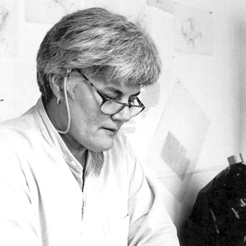 Black and white photo of a middle-aged woman with short grey hair and light skin. She sits against a wall and looks down, her glasses sliding down her nose slightly. She has an expression of concentration, as if she is focusing on something in her hands.