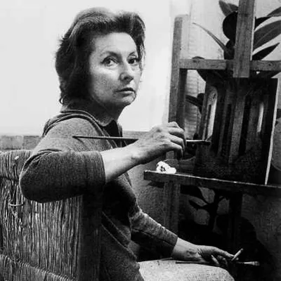 A black-and-white photograph of Remedios Varos, seated in front of an easel. She turns from an easel to face the camera, cigarette in her left hand and paintbrush in her right. Her brushed-back dark hair frames her light-skinned face, falling to just below her chin.