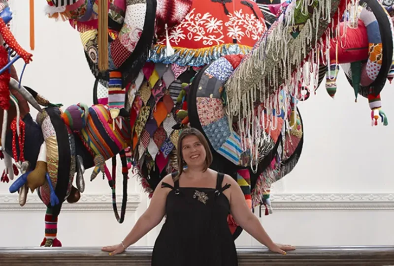 A light-skinned adult woman with bobbed brown hair wears a black sleeveless dress. She smiles wide and leans her hands back against a railing. Behind her is the lower portion of a large colorful fabric sculpture in front of a white wall.